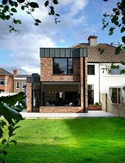 Rear view of a modern architect-designed house extension in Sandymount, Dublin with brick detailing and large open patio doors leading to a lush garden, under a bright sky with scattered clouds