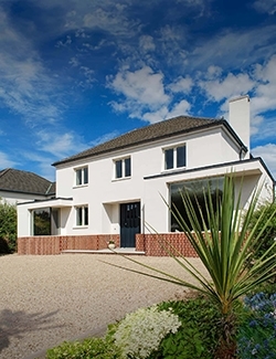 Two-story rebuilt white house in South Dublin with a textured architectural finish, fronted by a gravel driveway and vibrant green foliage under a sunny blue sky