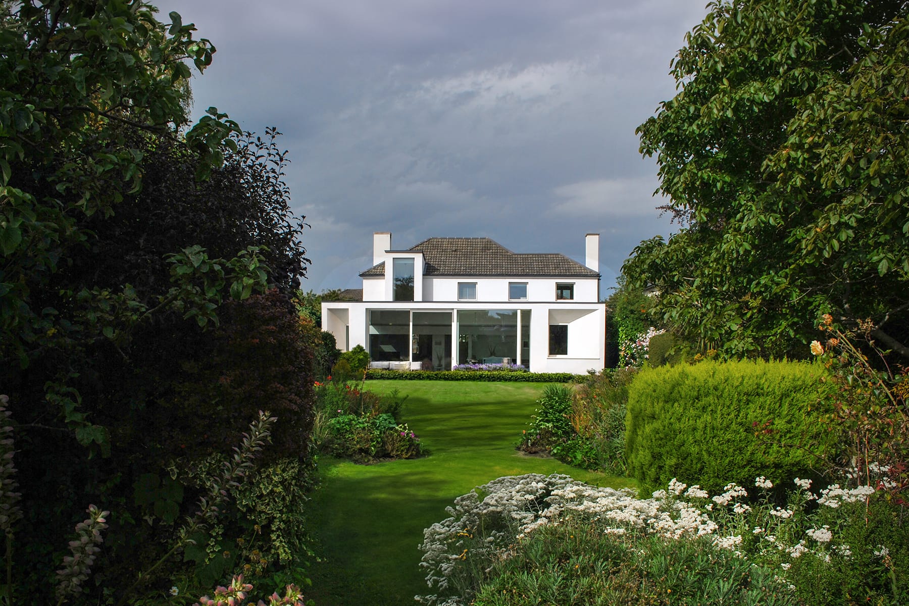 A spacious white house is designed by Dublin Architects, surrounded by lush greenery and trees in a beautifully landscaped garden